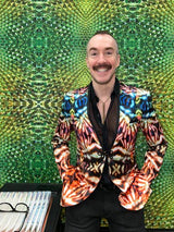 A smiling man with a mustache wearing a vibrant, multicolored patterned blazer over a black shirt, standing in front of a green textured kaleidoscope backdrop.