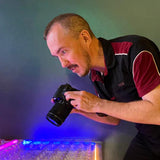 A man wearing a black and maroon polo shirt leans down to look through the viewfinder of a black DSLR camera, with blue and pink colored light reflecting off a glowing plastic surface below.