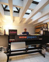 A large black offset printing press with 'wow papers' branding, shown in a workshop-like environment with a person standing behind it. The room has white ceiling beams, skylights, and tiled floors.
