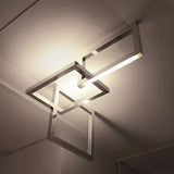 A modern geometric ceiling light fixture with intersecting square and rectangular metal frames, shown illuminated on a ceiling.
