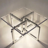 A modern geometric acrylic and metal illuminated table with a clear glass-like top and intersecting silver metal frame legs, photographed in a minimalist studio setting.