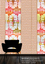 A wallpaper sample display featuring vertical stripes with a textured rose gold and beige background, overlaid with three vertical panels of an abstract geometric pattern in coral, yellow, and olive green. A black chair silhouette is shown at the bottom against a dark wood floor, with a 'wowpapers' brand watermark at the bottom right.