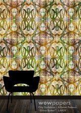 A large-scale wallpaper pattern featuring an abstract, vintage-style allover design in olive green, gold, beige, and brown tones. A black silhouette of a modern armchair is shown in the foreground against the patterned wall, with a dark wood floor at the bottom.
